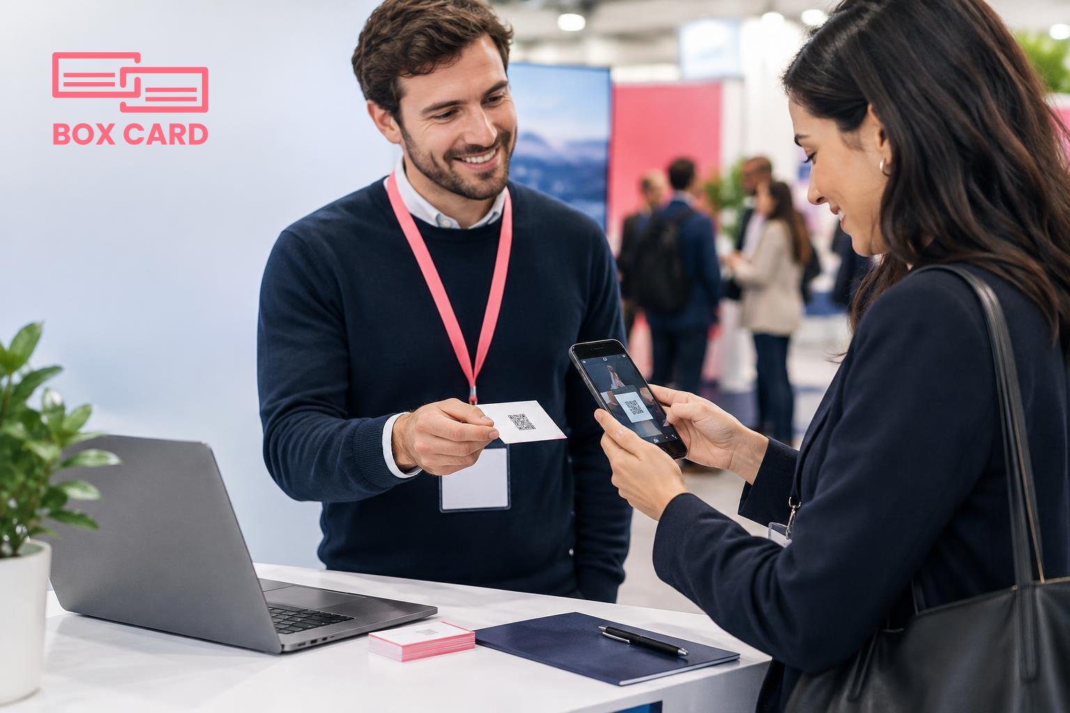 Two professionals exchanging business cards at a conference booth while one scans the other card with a smartphone, name badges and a laptop on the table, editorial photograph