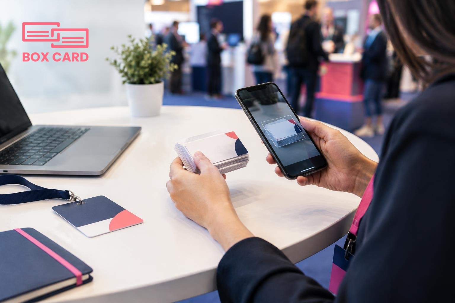 A conference attendee scanning a stack of paper business cards with a smartphone, with a conference badge and a laptop on a table, editorial photograph