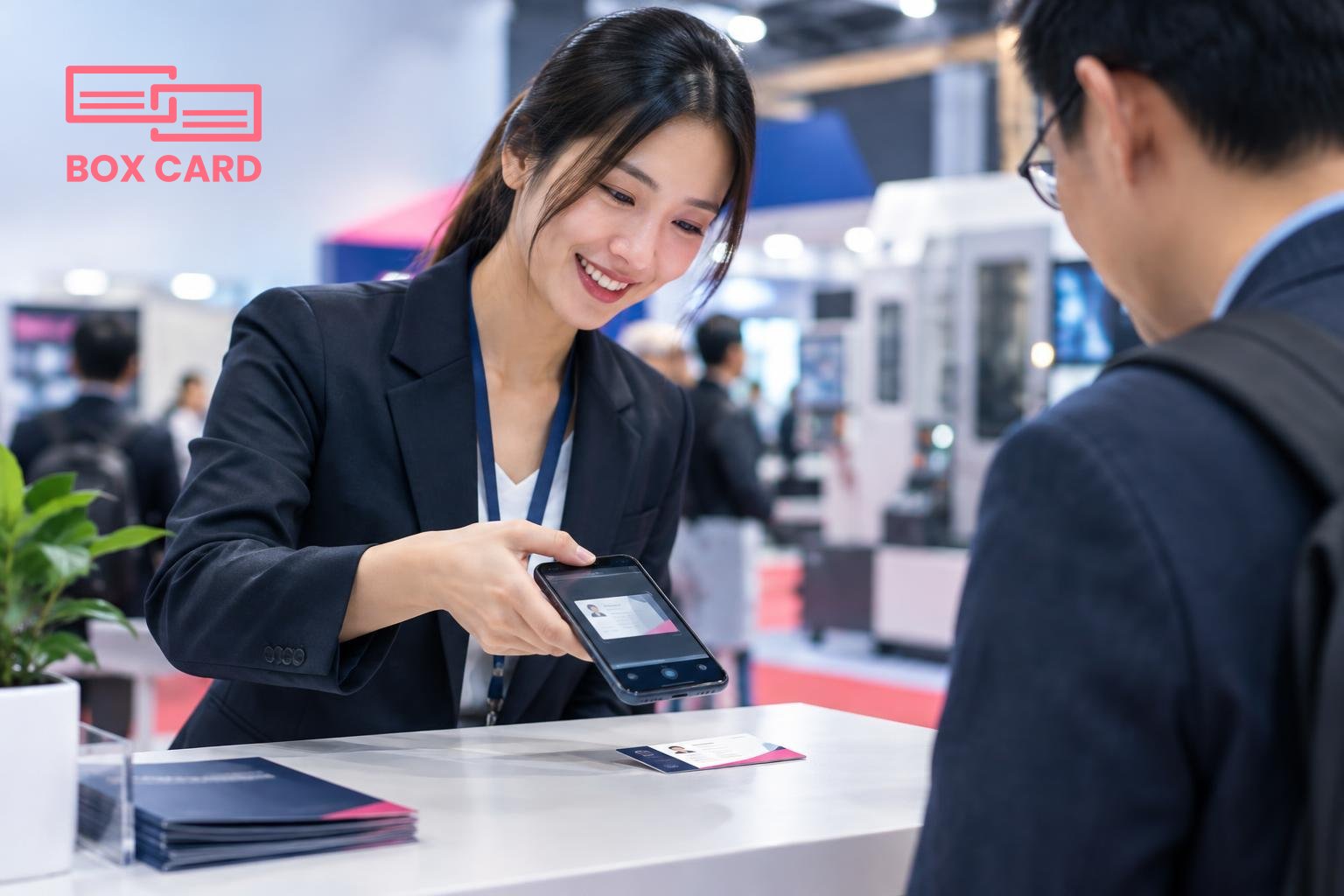 Một nhân viên trao đổi với khách trước gian hàng, đang dùng điện thoại quét danh thiếp trên bàn, màn hình điện thoại hiển thị giao diện ứng dụng Boxcard với thẻ liên hệ và ghi chú, ảnh phong cách edit